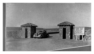 A black and white photo of an old car parked in front of two stone gates.